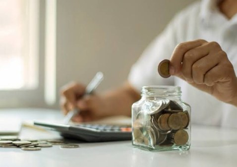 Hand die een muntje in een spaarpot met munten laat vallen, met op de achtergrond iemand die een rekenmachine gebruikt
